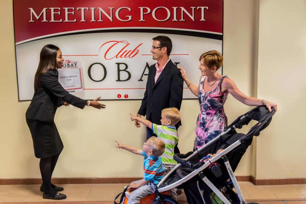 Woman welcoming family with two kids and stroller at indoor "Meeting Point" sign in Negril Jamaica.
