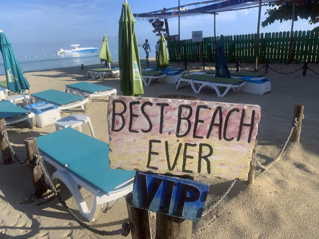 Negril Jamaica beach with lounge chairs, umbrellas, and a "BEST BEACH EVER VIP" sign by the shoreline.