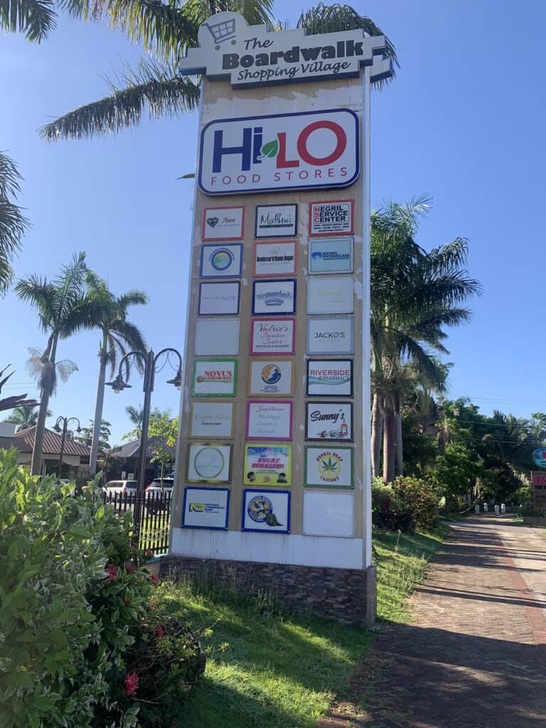 Boardwalk Shopping Village sign with shops listed, surrounded by palm trees in Negril Jamaica.