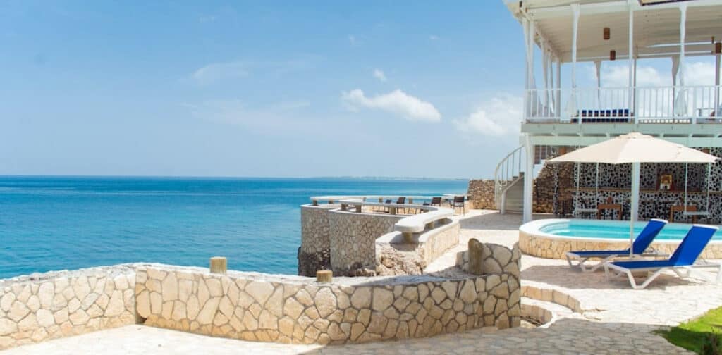 Negril Jamaica stone patio with pool, lounge chairs, and umbrella by the ocean near a white seaside building.