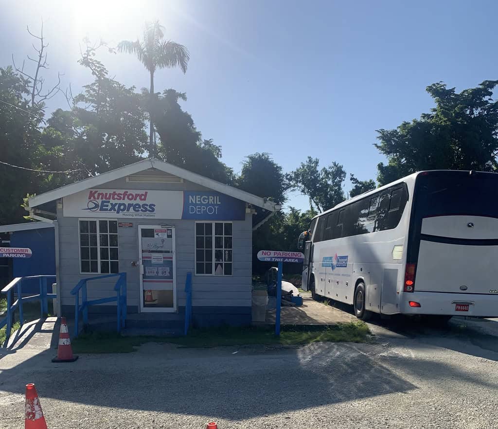 Knutsford Express Negril Jamaica depot with white bus parked outside, trees in background, sunny weather.