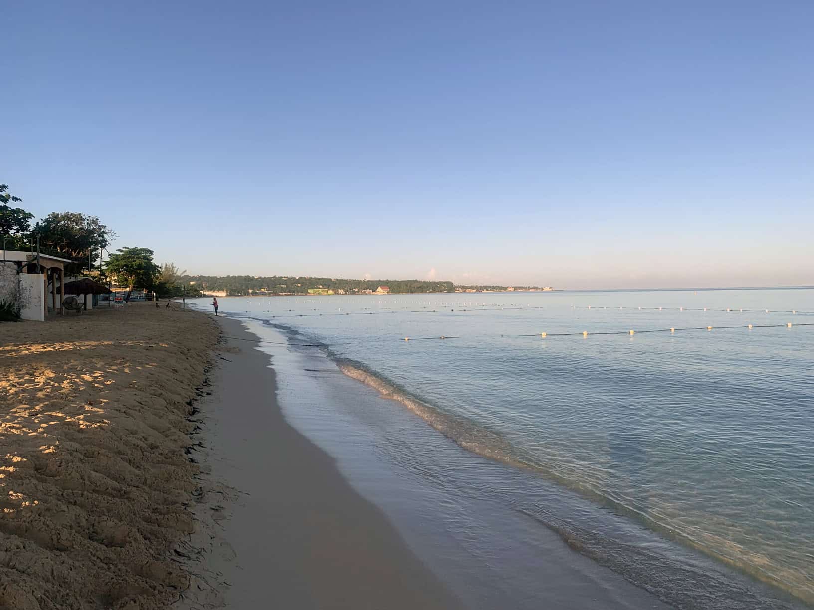 Negril Jamaica beach in early morning, gentle waves on sandy shore, clear sky, and trees lining the left side.