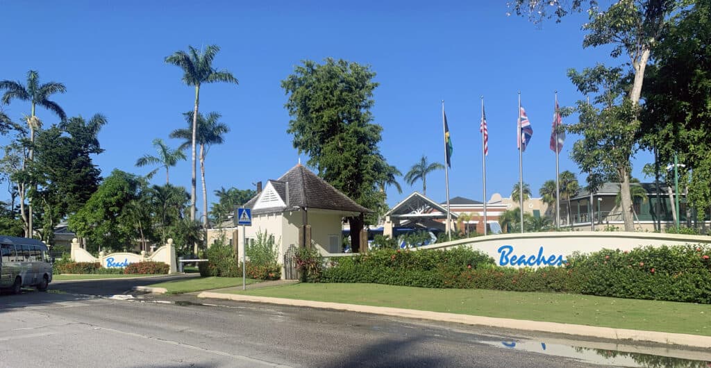 Beaches resort entrance in Negril Jamaica with flags, palm trees, green lawn, and blue sky in the background.