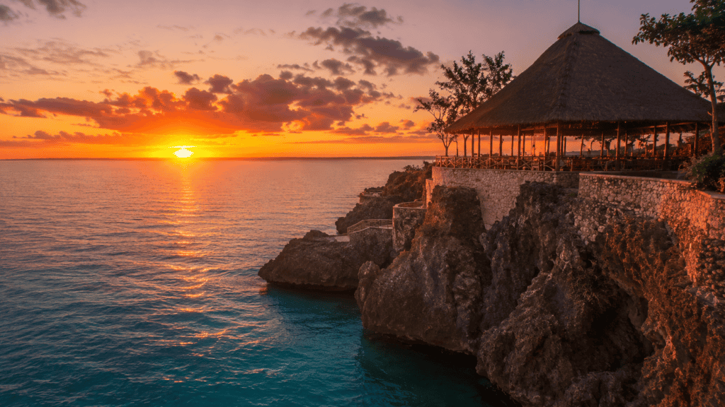 Negril Jamaica sunset with thatched-roof pavilion on rocky cliffs overlooking ocean and colorful sky.