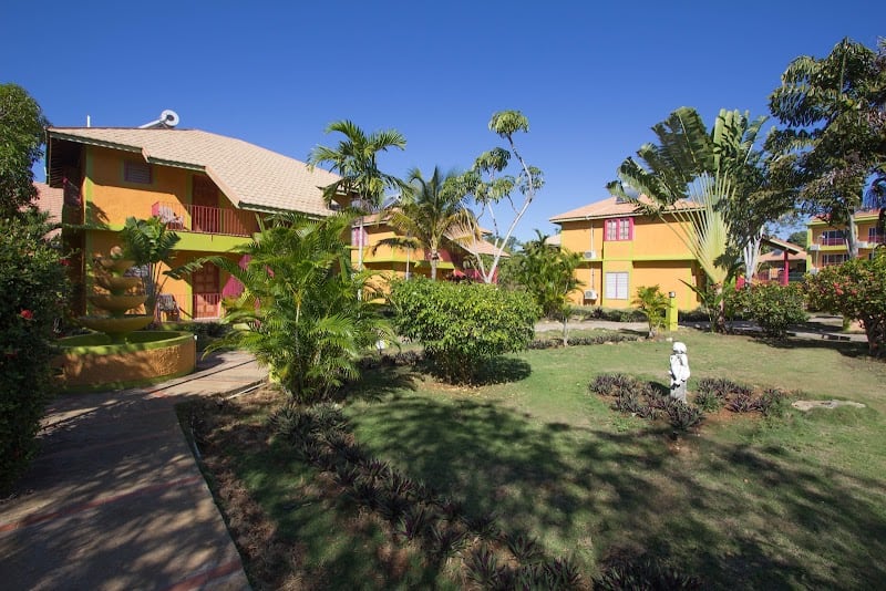 Colorful tropical buildings with red trim, palm trees, and gardens in Negril Jamaica under a clear blue sky.