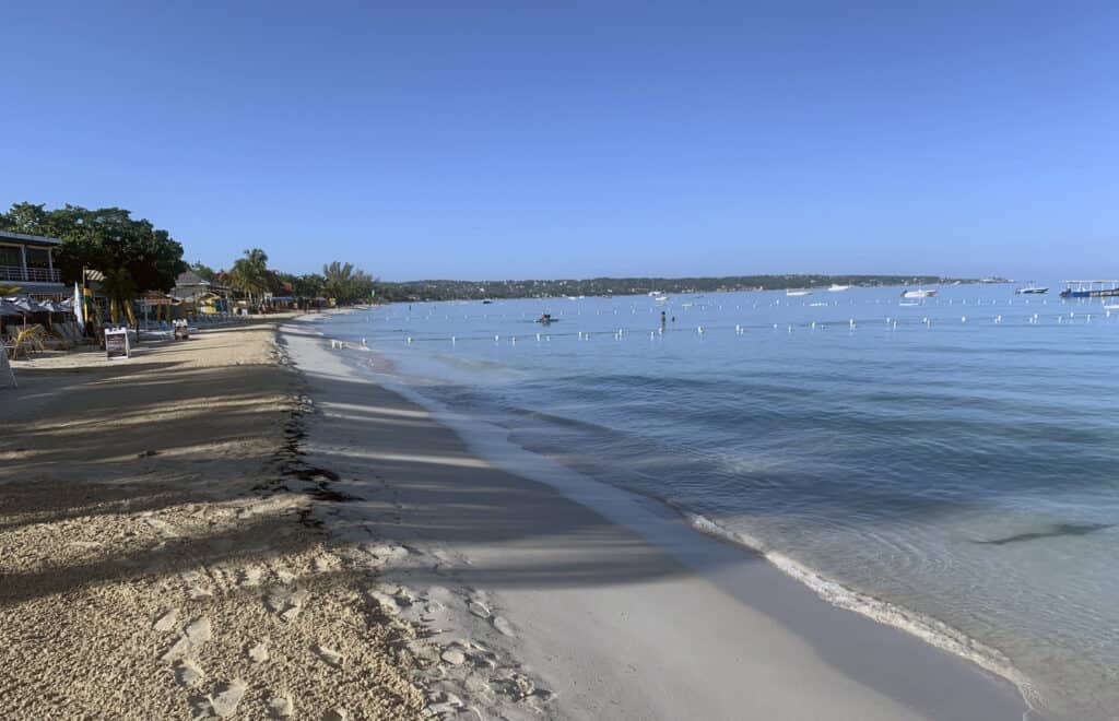 Negril Jamaica sandy beach with calm blue water, boats, and shoreline buildings under a clear sky.