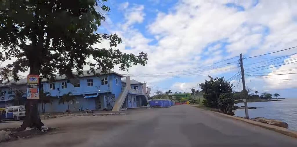 Curved street by blue buildings and waterfront in Negril Jamaica under a partly cloudy sky.