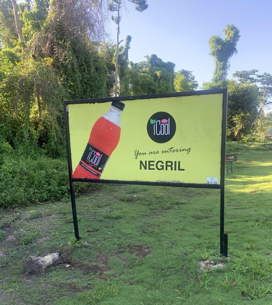 Yellow welcome sign with red drink bottle reading "You're entering Negril," surrounded by lush greenery in Negril, Jamaica.